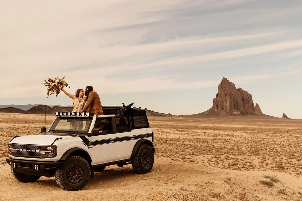 A couple in wedding attire standing in a white off-road vehicle with a mountain in the desert behind them.