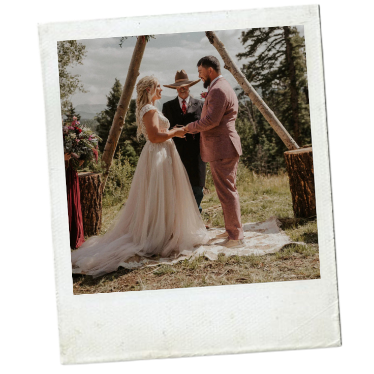 Bride and groom holding hands during their outdoor wedding ceremony, officiant in the background, surrounded by trees and logs, in a natural setting.