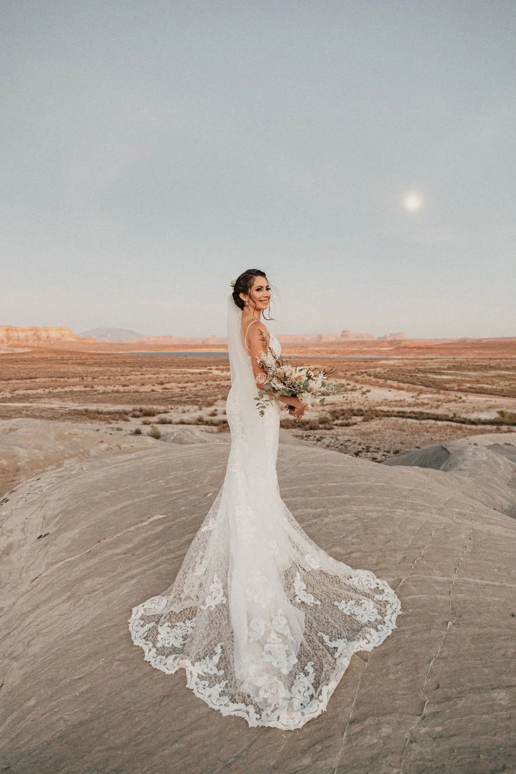 Bridal portrait of a woman in a white lace wedding dress holding a bouquet, standing on a rock formation in a desert landscape with distant mesas and a bright sky.