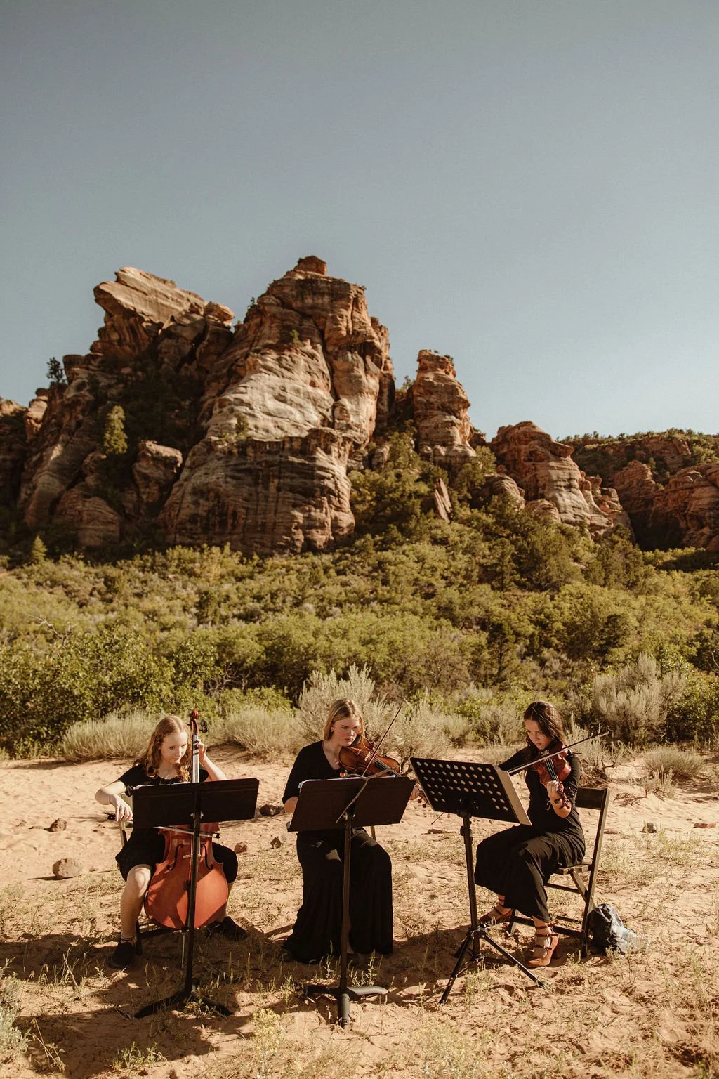 Three women playing string instruments outdoors in a desert-like area with large rocky formations and green bushes.