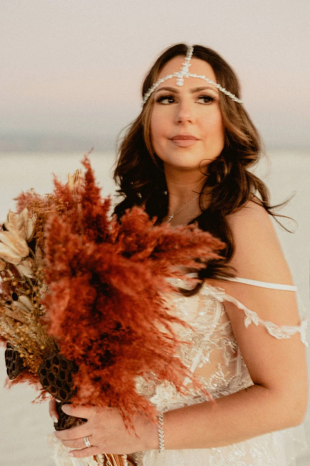 A woman with long brown hair and light makeup, wearing a white lace dress and a pearl headband, holding a bouquet of dried flowers, outdoors during sunset.