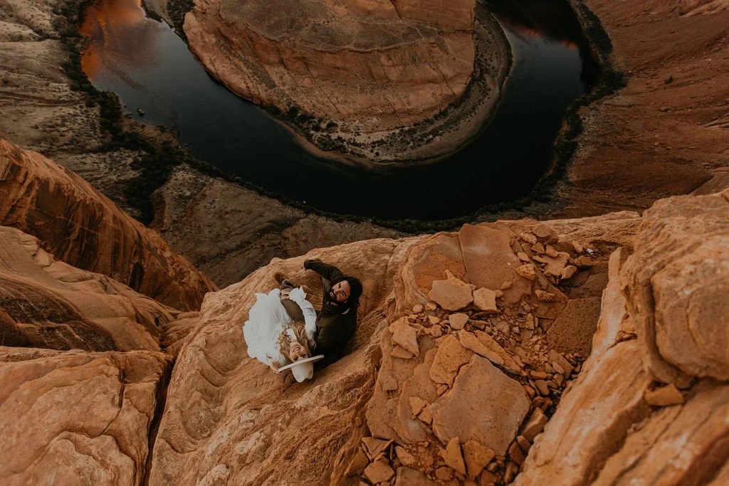 A couple dressed in wedding attire sits on a rocky ledge overlooking a winding river in a canyon.