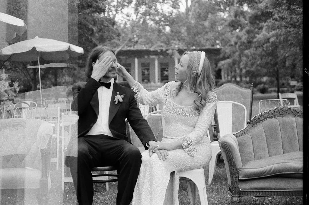 A woman dressed as a bride playfully touching the forehead of a man dressed as a groom, who is smiling with eyes closed, at an outdoor wedding reception with chairs, tables, and trees in the background.