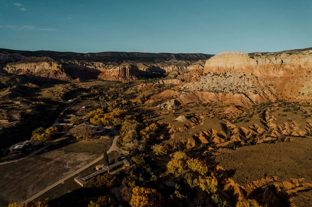 Aerial view of a landscape with layered red, orange, and beige rock formations, sparse vegetation, and a few buildings amidst trees with green and yellow leaves.