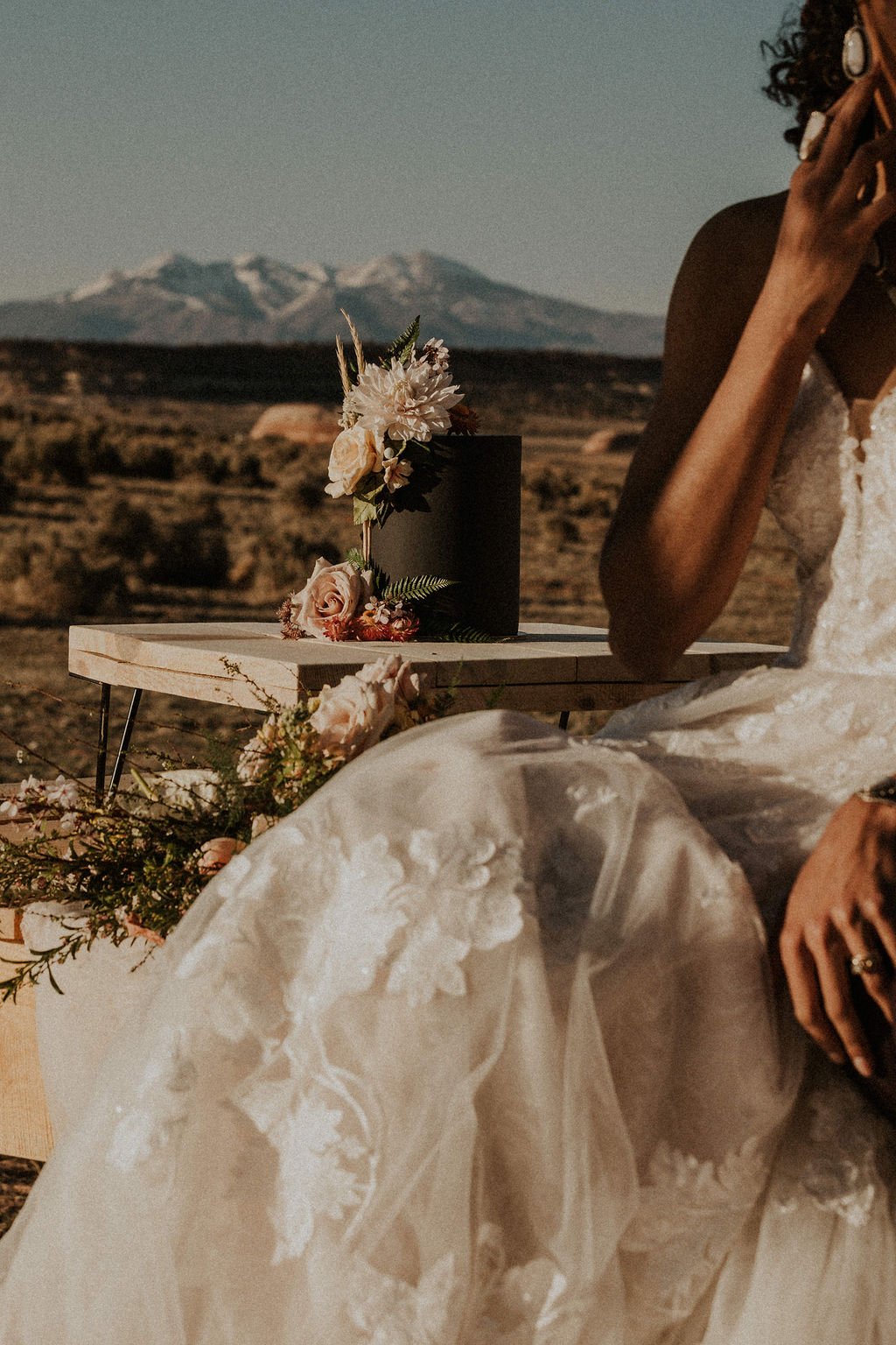 A bride in a white lace wedding dress sitting outdoors with a small wooden table holding a black wedding cake decorated with flowers, set against a mountainous landscape.
