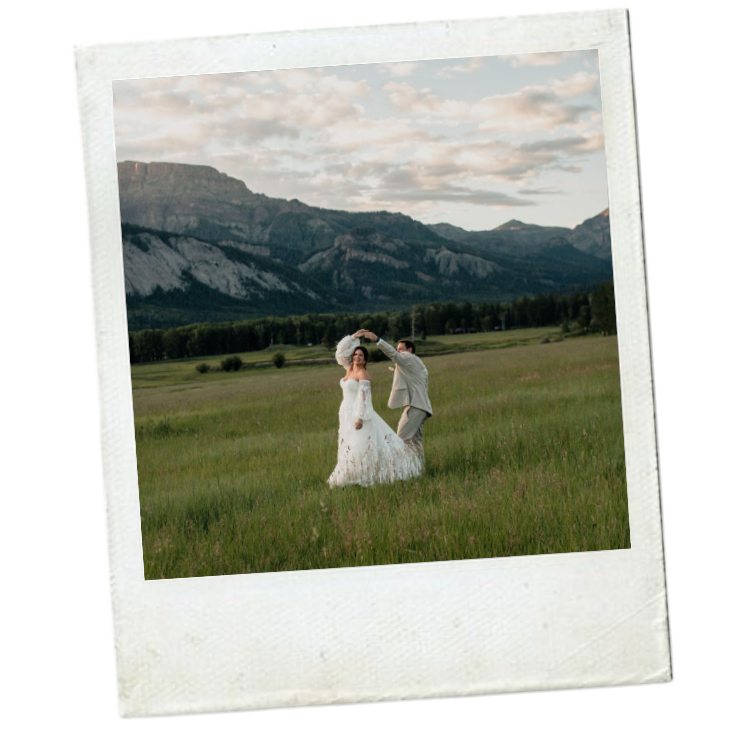 A couple dancing in a grassy field with a mountain range in the background, dressed in wedding attire during sunset.