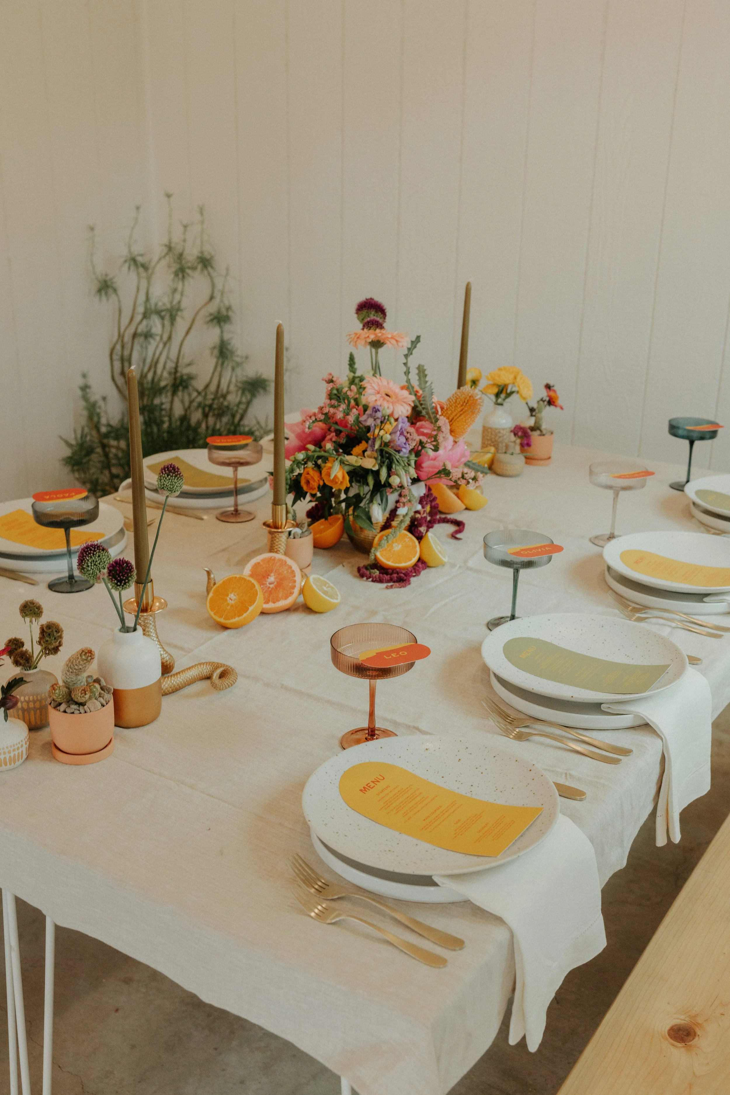 A decorated dining table with plates, gold-colored cutlery, and colorful drink glasses. The table has a beige tablecloth, and a centerpiece featuring various flowers and fruit slices, including oranges and lemons. There are also small pots with plant
