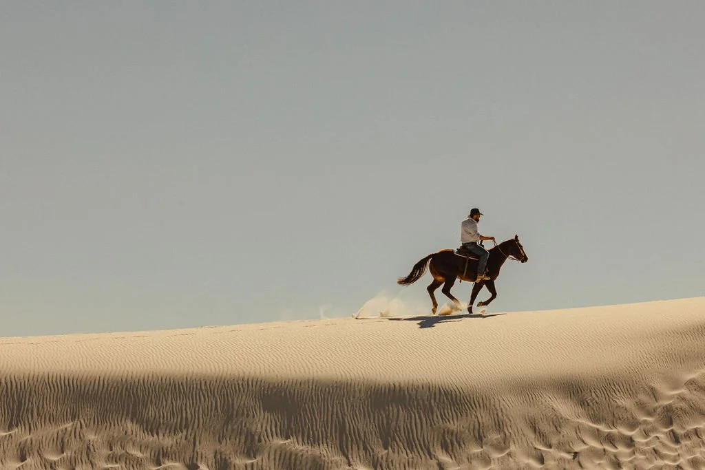 A person riding a horse across desert sand dunes under a clear sky.