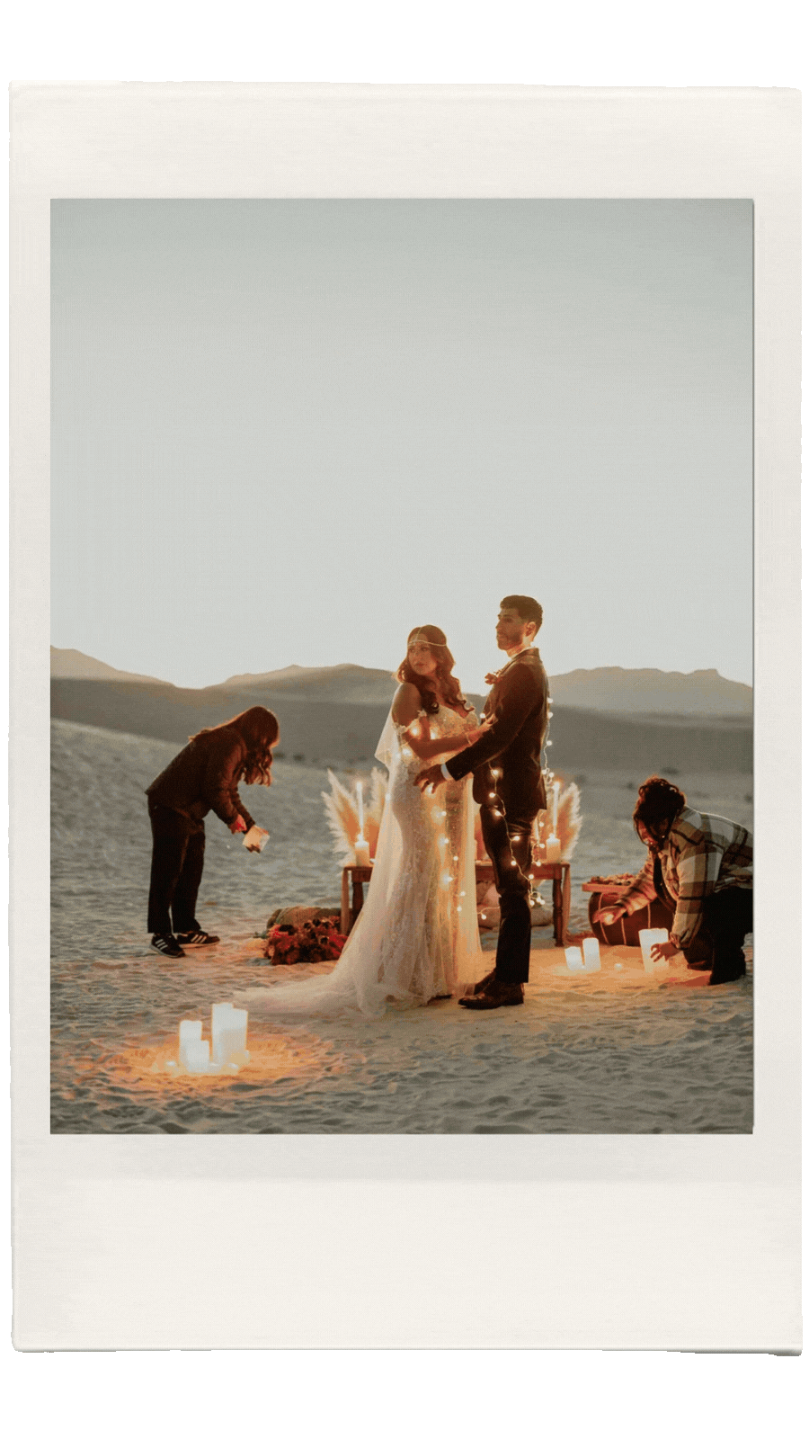 A couple in wedding attire holding hands during an outdoor wedding on a beach at sunset, surrounded by floating candles and decorative flowers.