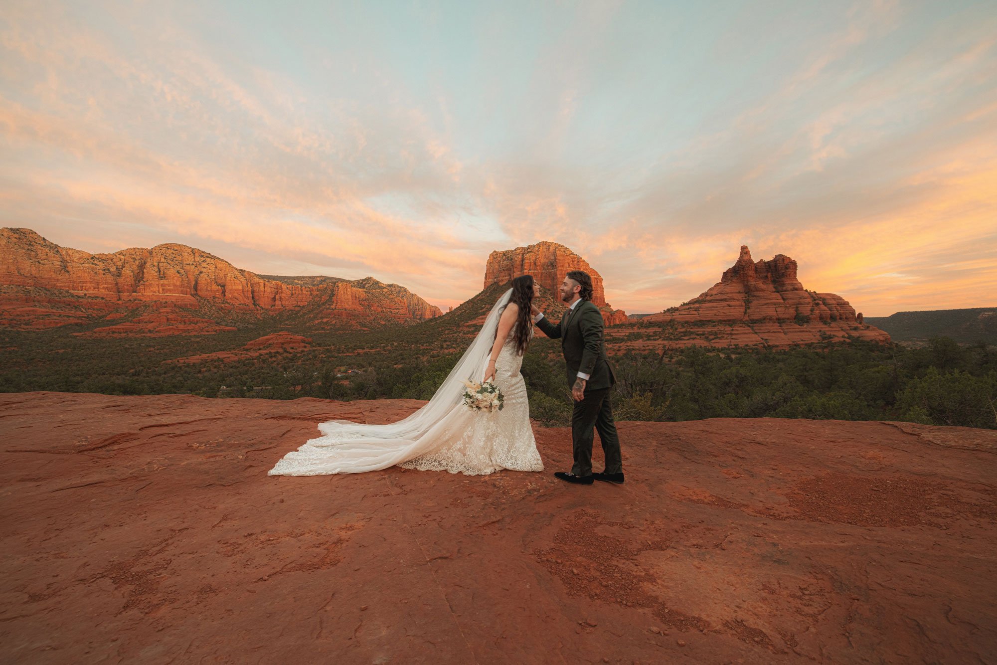 A bride and groom standing on a red rock in front of a desert mountain range in Arizona at sunset. The bride, in a white lace wedding dress and veil, holds a bouquet and the groom, in a dark suit, is touching her face.