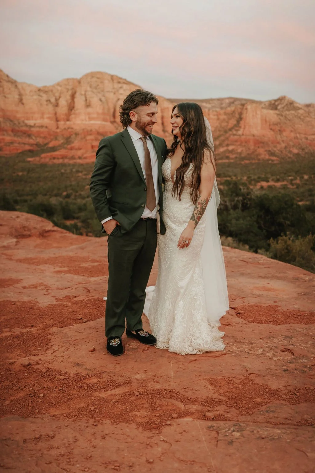 A bride and groom standing on a red rock formation at sunset with mountains in the background, smiling at each other.