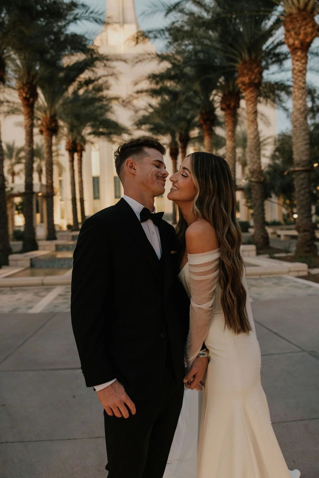 A couple dressed in formal wedding attire, a man in a black tuxedo and a woman in a white wedding gown, standing close together outdoors with palm trees and a building in the background during sunset.