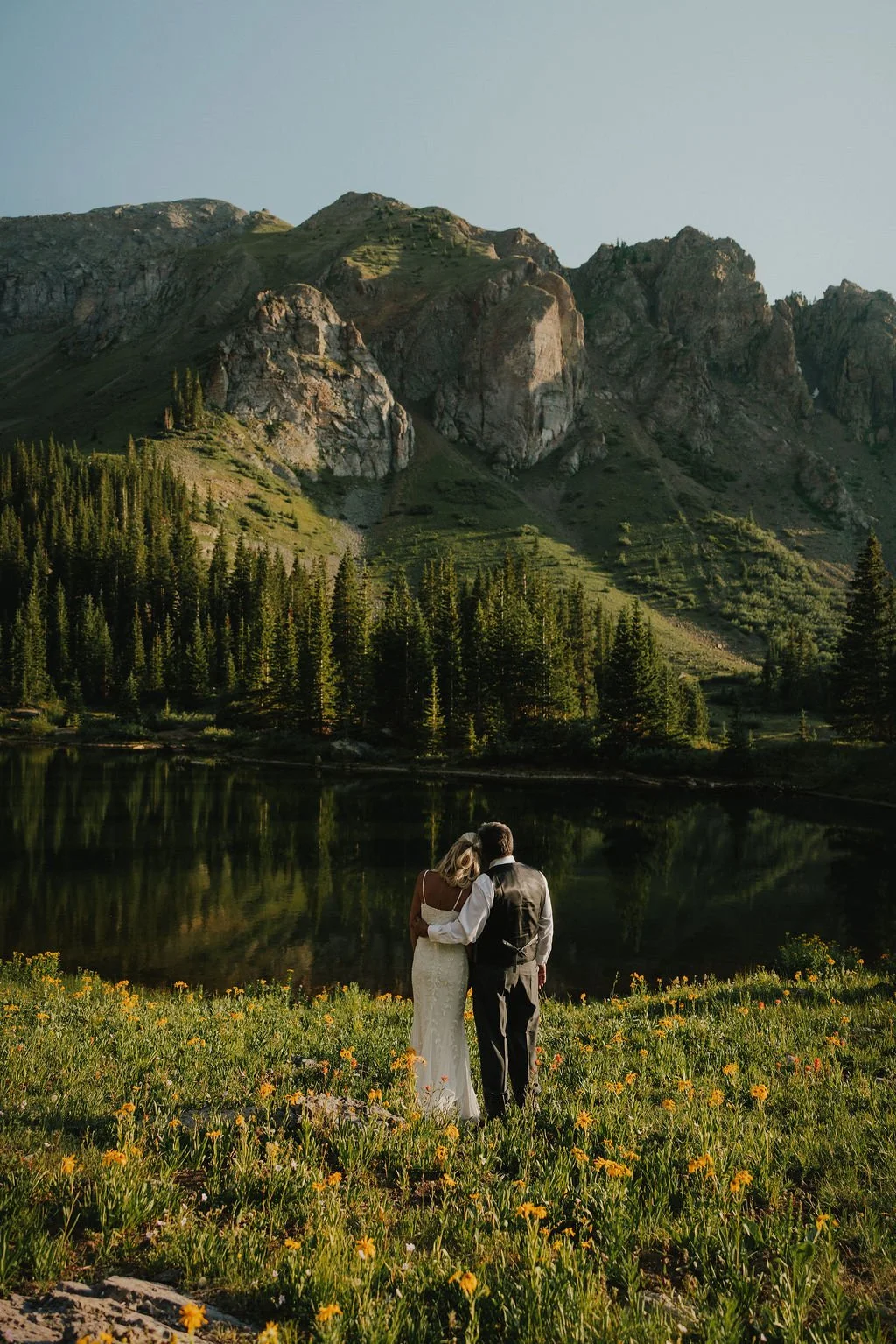 A couple stands close together, facing a mountain lake surrounded by tall evergreen trees and rugged mountains, during sunset.