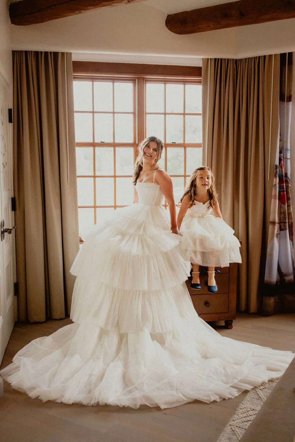A woman and a young girl in wedding dresses standing inside a room near a large window with beige curtains, with both smiling and the woman lifting the skirt of her dress.