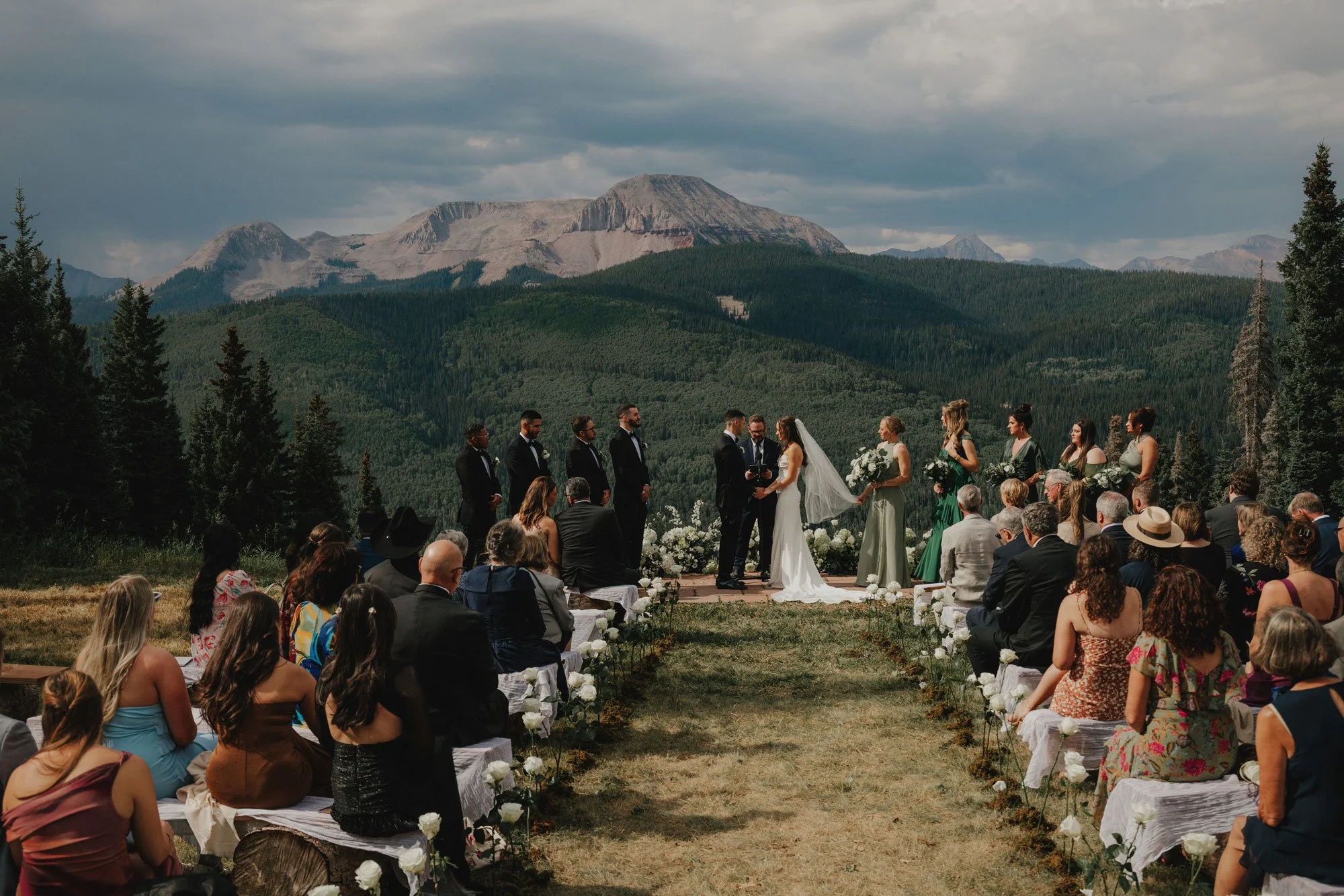 Outdoor mountain wedding ceremony with bride, groom, officiant, bridesmaids, and groomsmen, surrounded by trees and mountains under cloudy sky.