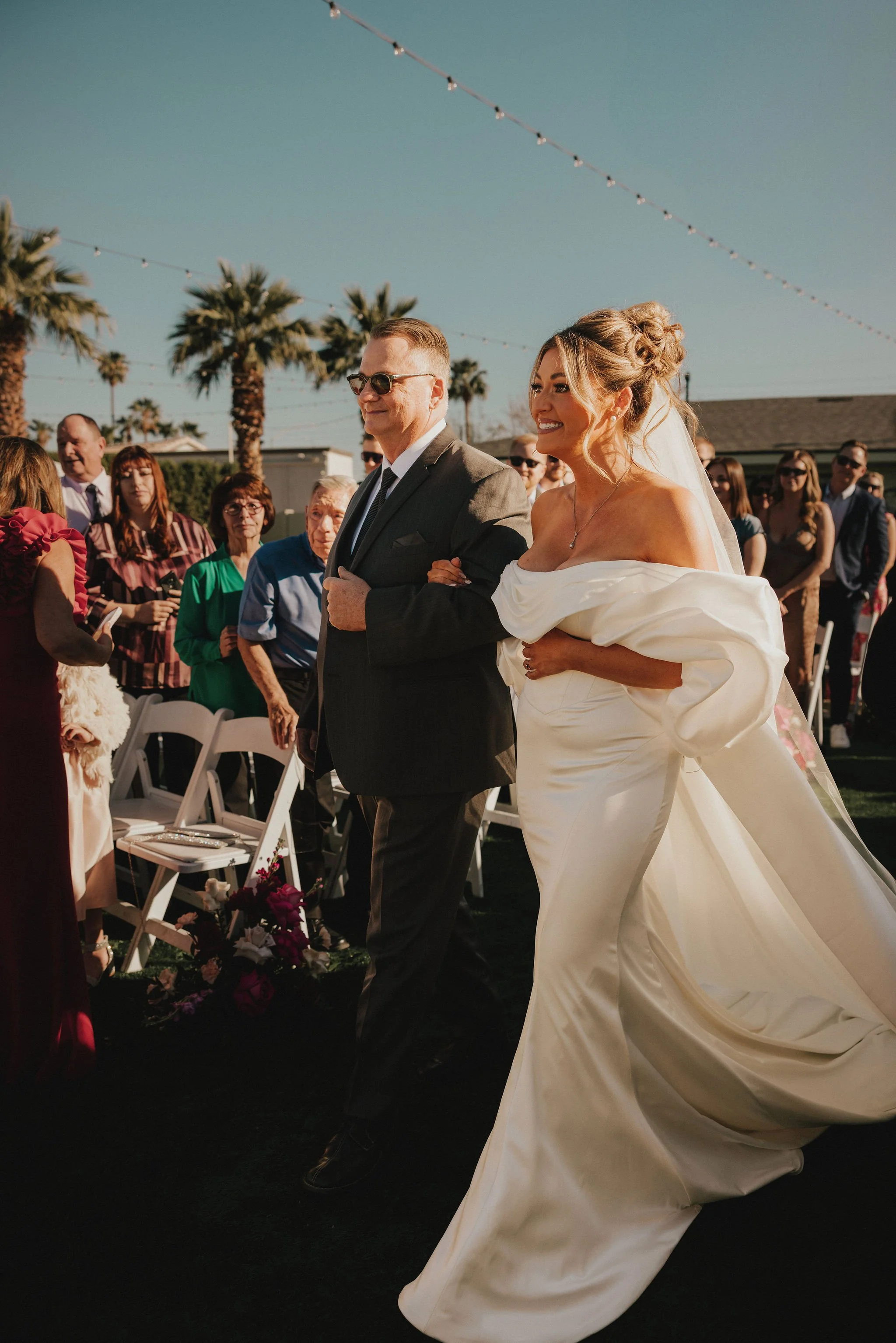 A bride in a white off-shoulder wedding gown walking down the aisle alongside a man in a gray suit at an outdoor wedding ceremony.