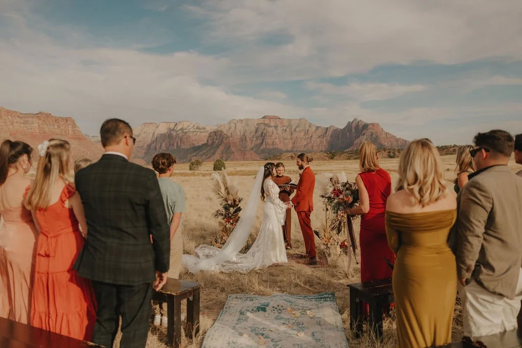 A couple getting married outdoors in a desert with red rock formations in the background, surrounded by friends and family.