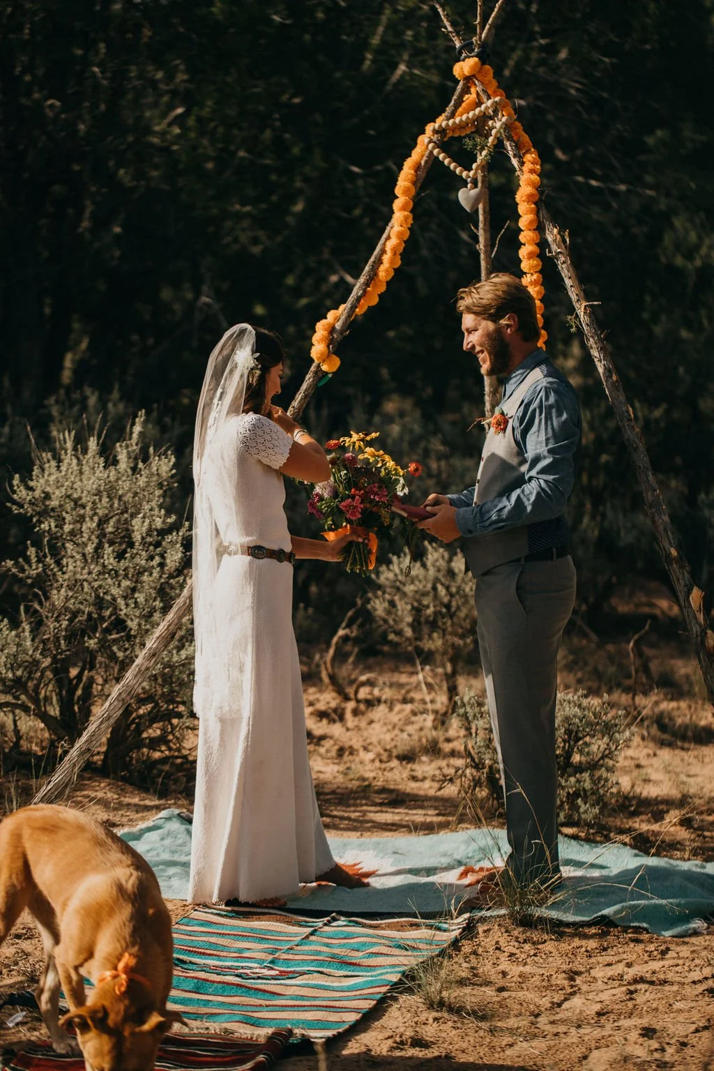 A couple exchanges wedding vows outdoors under a rustic wooden arch decorated with orange marigold flowers, with a bride in a white dress and veil holding a bouquet, and a groom in a blue shirt and gray vest smiling. A dog is visible at the bottom, s