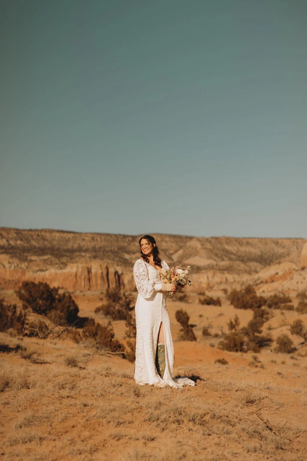 A woman in a white lace dress holding a bouquet of flowers standing in a desert landscape with rock formations and sparse vegetation under a clear blue sky.