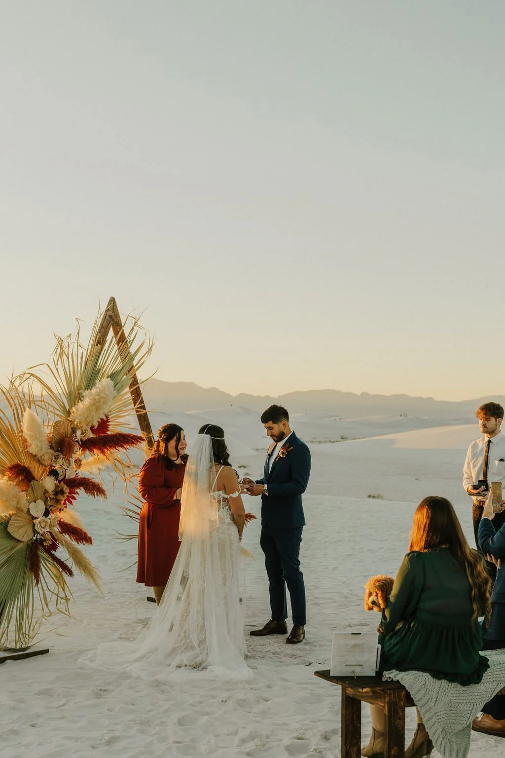 A wedding ceremony taking place on a sandy beach at sunset. A couple is exchanging rings in front of a floral arch with dried palm leaves and flowers. Guests are watching, and the bride is wearing a white lace dress with a veil, while the groom is in