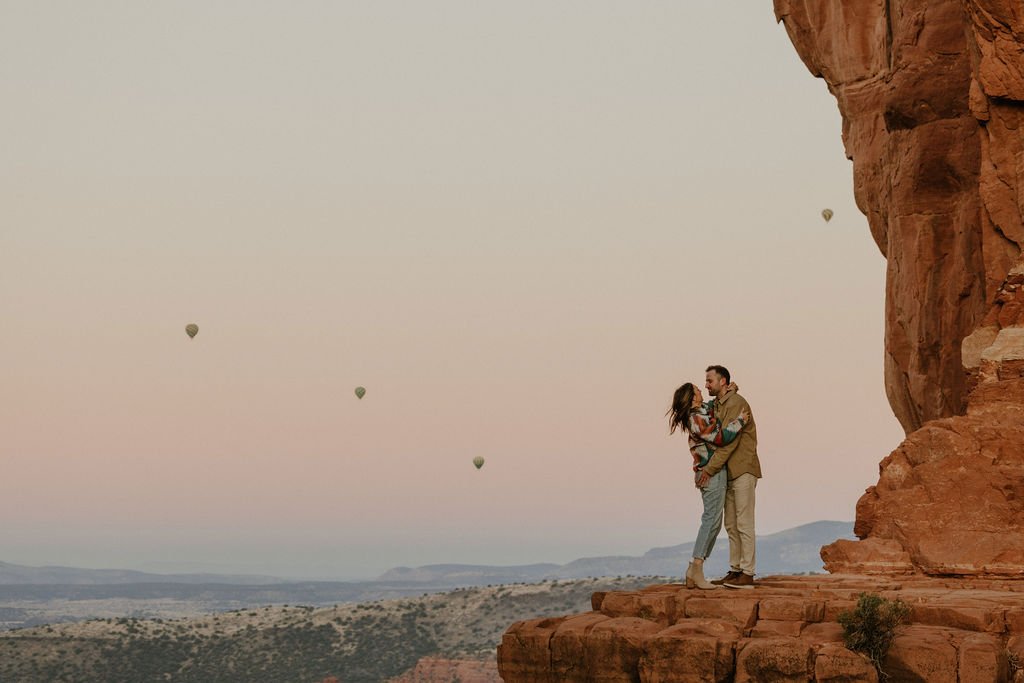 A couple standing on a rocky ledge, embracing and smiling at each other against a desert landscape with a few hot air balloons in the sky at sunrise or sunset.