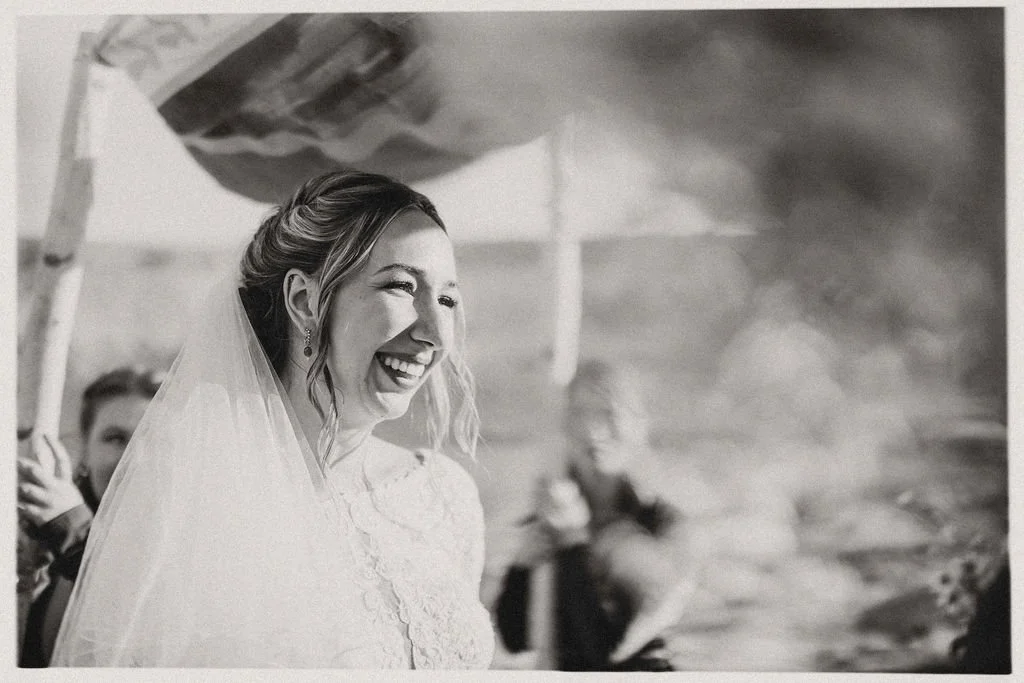 Black and white photo of a smiling bride with a veil, standing outdoors at her wedding, with friends visible in the background.