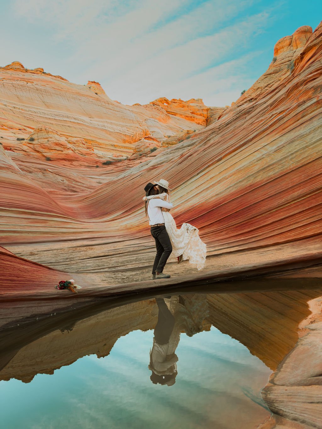 A couple dancing in a canyon with colorful layered rock formations, one person lifting the other, and their reflection visible in a pool of water at the canyon floor.