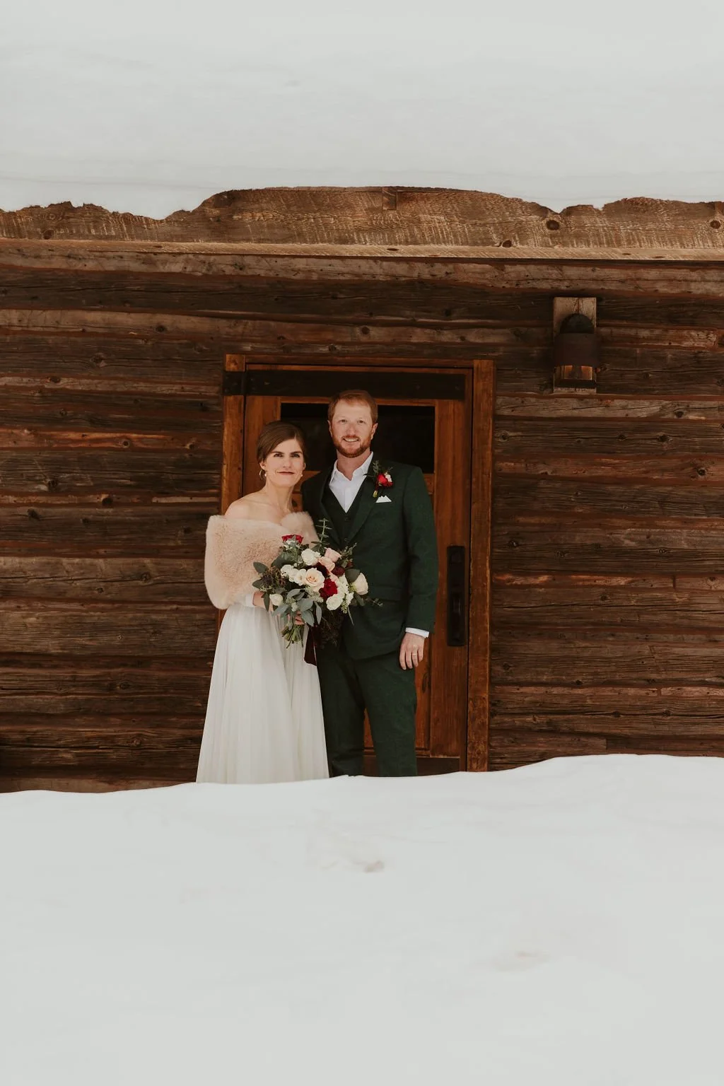 A bride and groom in wedding attire standing outside a wooden cabin in the snow, holding a bouquet of flowers.