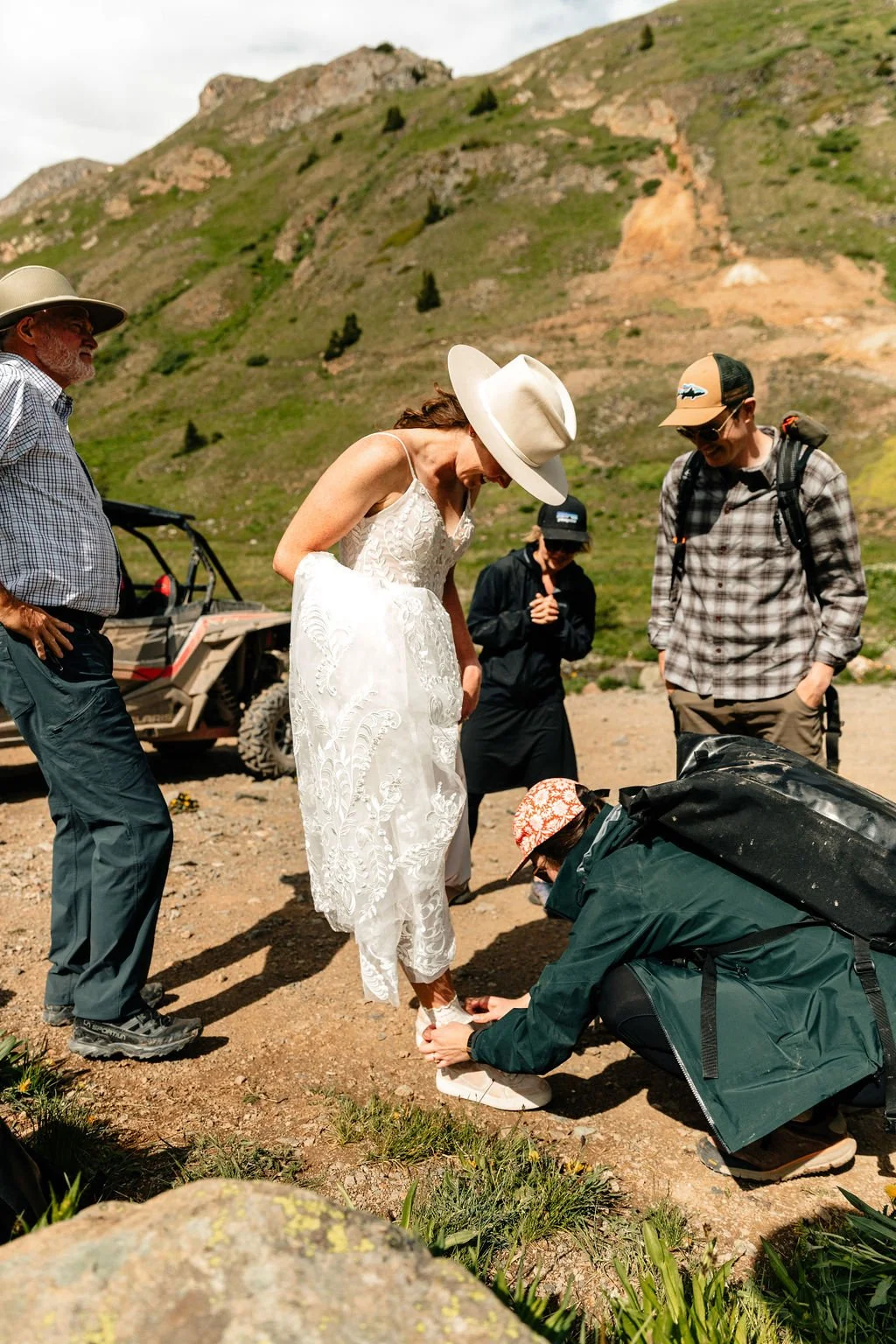 A woman in a white dress and wide-brimmed hat having her shoe tied in an outdoor mountainous area, with several people around her, some wearing hats, one with a backpack, and an off-road vehicle in the background.