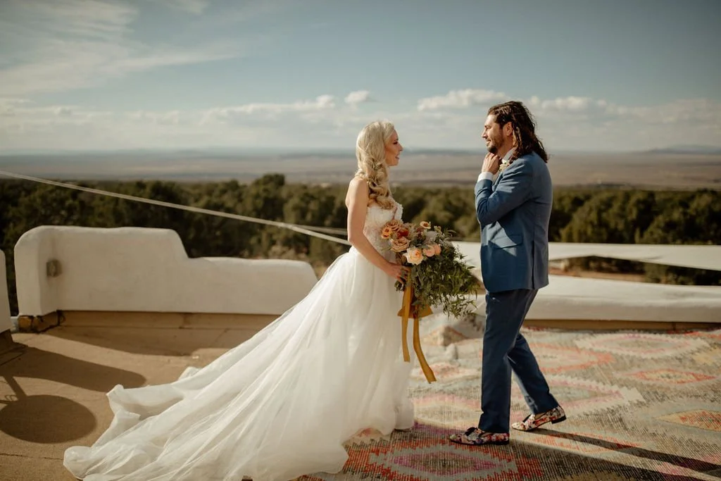Bride and groom exchanging vows on a rooftop with scenic landscape in the background, the bride in a white wedding gown holding a bouquet, the groom in a blue suit.