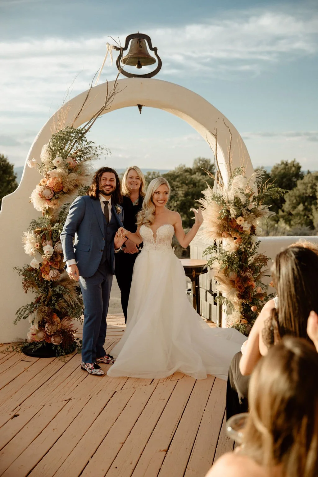 A wedding ceremony taking place on an outdoor wooden deck under a white arch decorated with flowers, with the bride and groom holding hands and smiling, surrounded by an audience, during sunset.