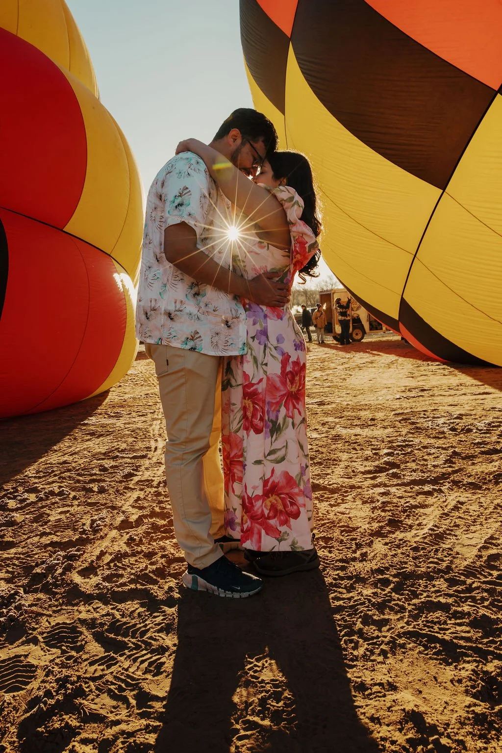 A couple embracing at a hot air balloon event during sunset, with large colorful balloons around them and photographers in the background.