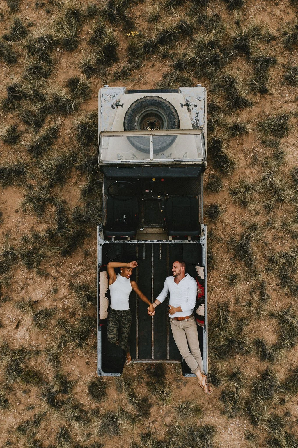 A man and a woman lying in the back of an open truck in a desert area, holding hands, and smiling at each other.