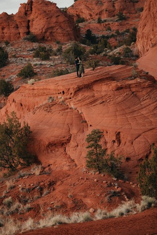 A person standing on a large, red rock formation in a desert landscape with red cliffs and sparse vegetation.