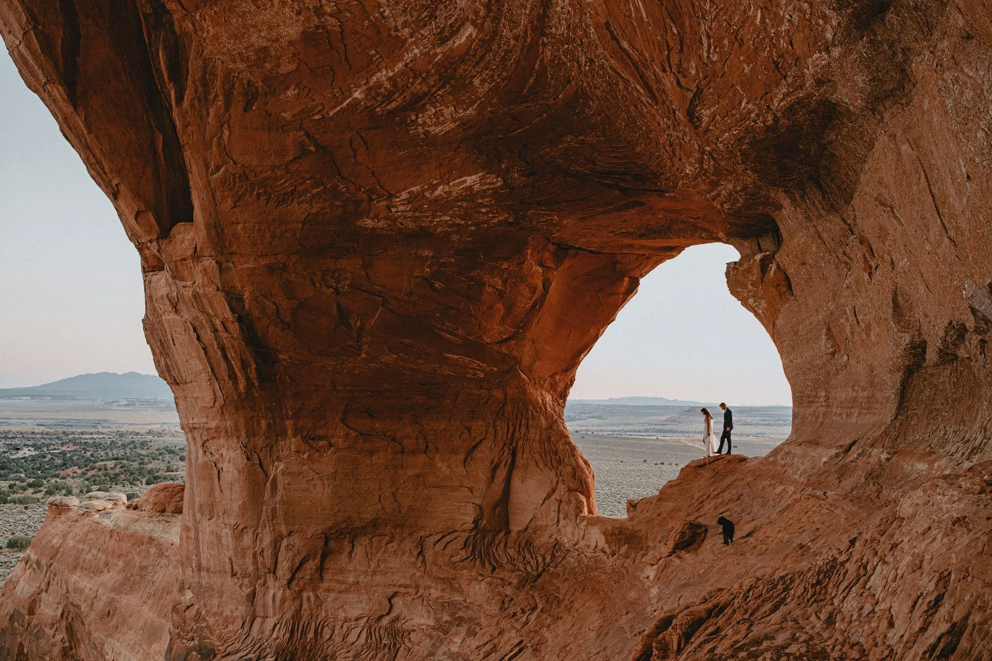 A newly wed couple holding hands and walking on a rocky cliff within a large red sandstone arch formation in the Utah desert.