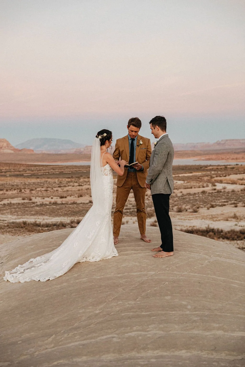 Bride and groom standing barefoot on a desert rock at sunset, exchanging vows with officiant reading from a book.