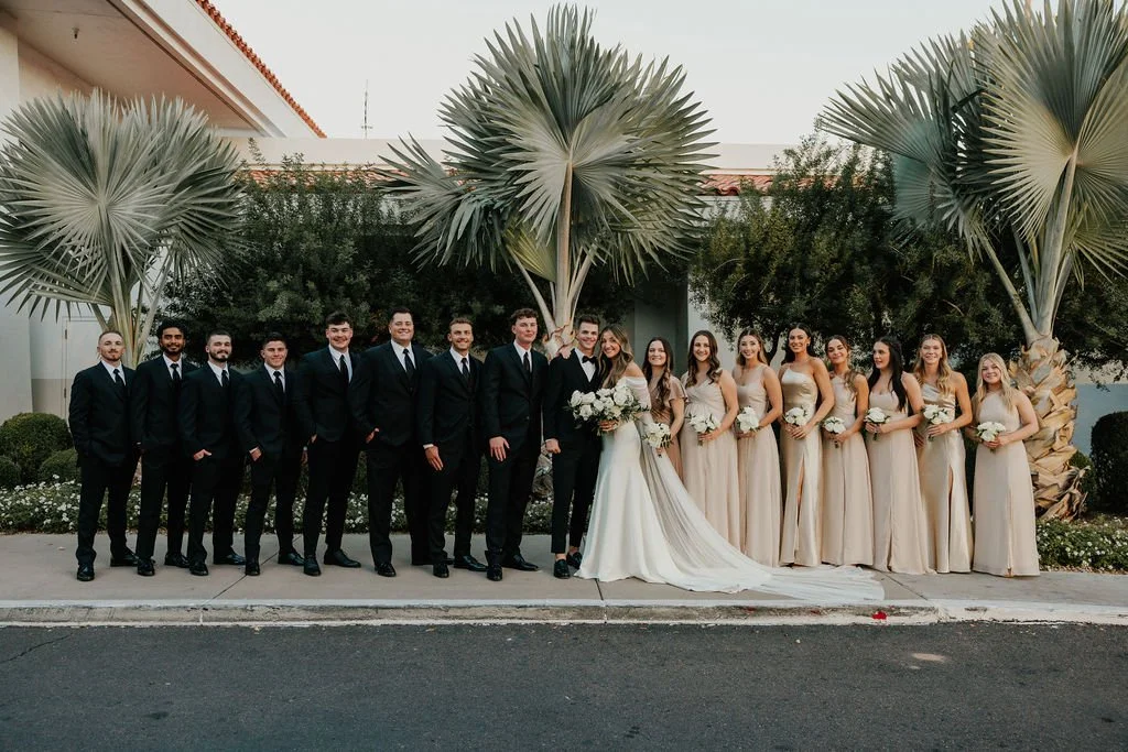 A wedding party with 14 people, including bridesmaids in beige dresses holding bouquets, groomsmen in black suits and ties, and a bride in a white gown and veil holding a bouquet, standing outdoors in front of palm trees and greenery.