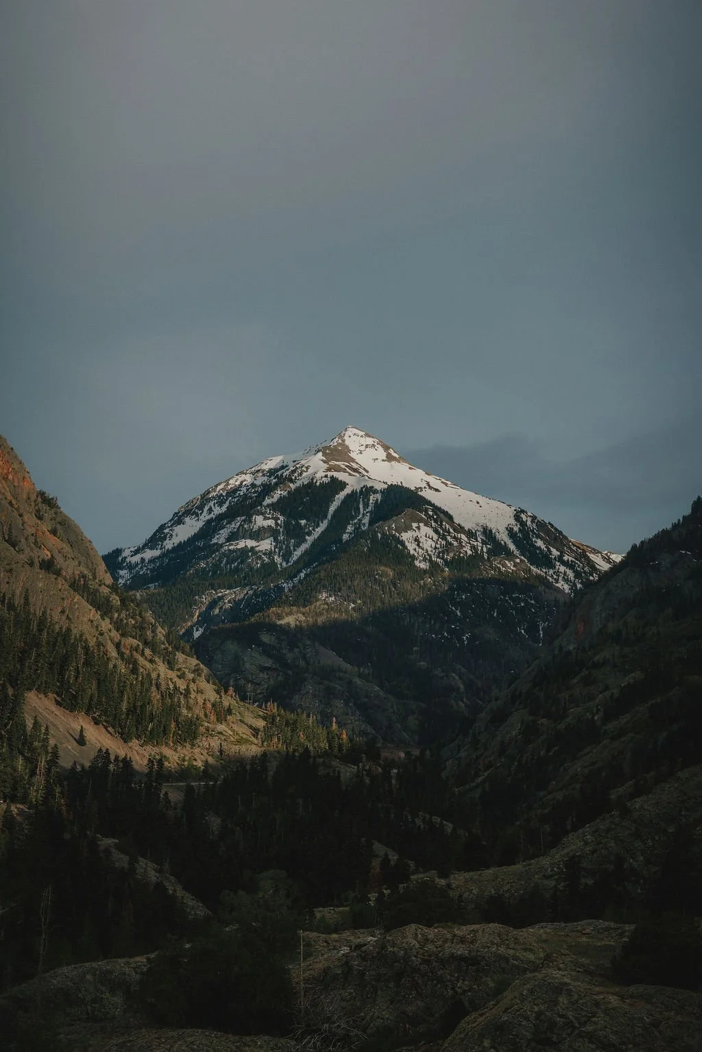 Snow-capped mountain surrounded by forested slopes under cloudy sky.