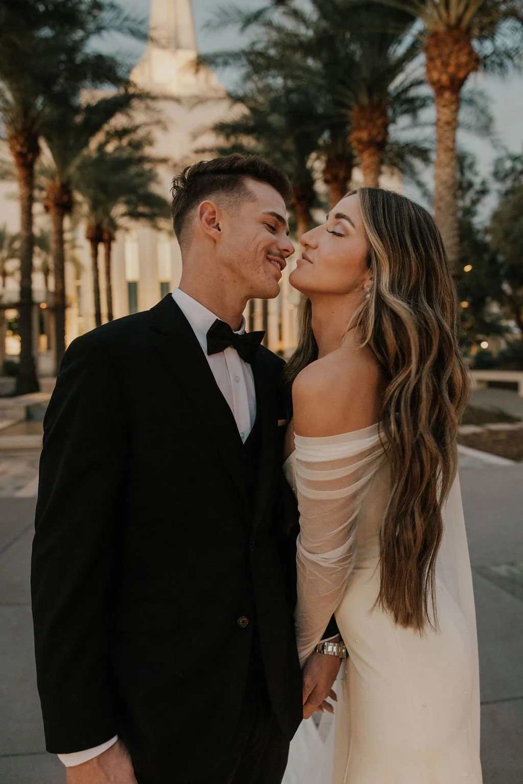A couple dressed in wedding attire standing close together outdoors with tall palm trees and a building in the background, likely during sunset.