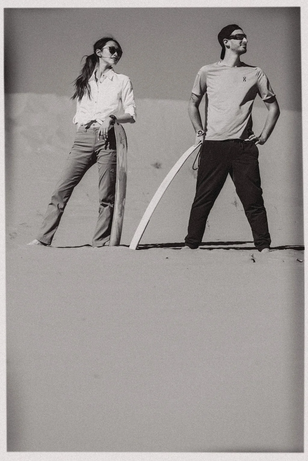 A man and woman standing on a sandy beach with a long surfboard. Both are wearing sunglasses and casual clothing, smiling and posing confidently.