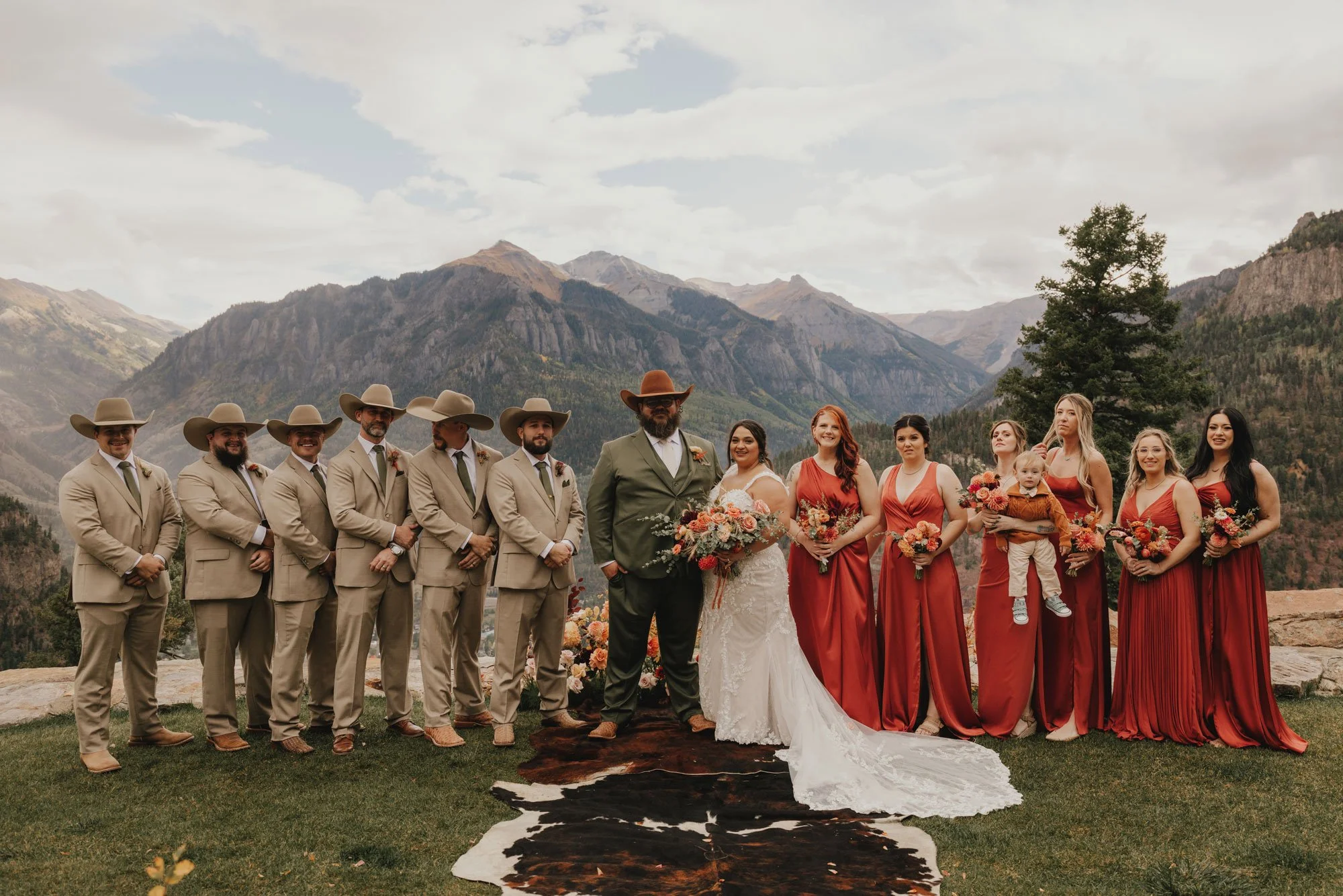 Group of wedding party members, including bridesmaids in red dresses and groomsmen in beige suits with cowboy hats, standing outdoors on a grassy area with mountains in the background. The bride and groom are in the center, with the bride in a white 