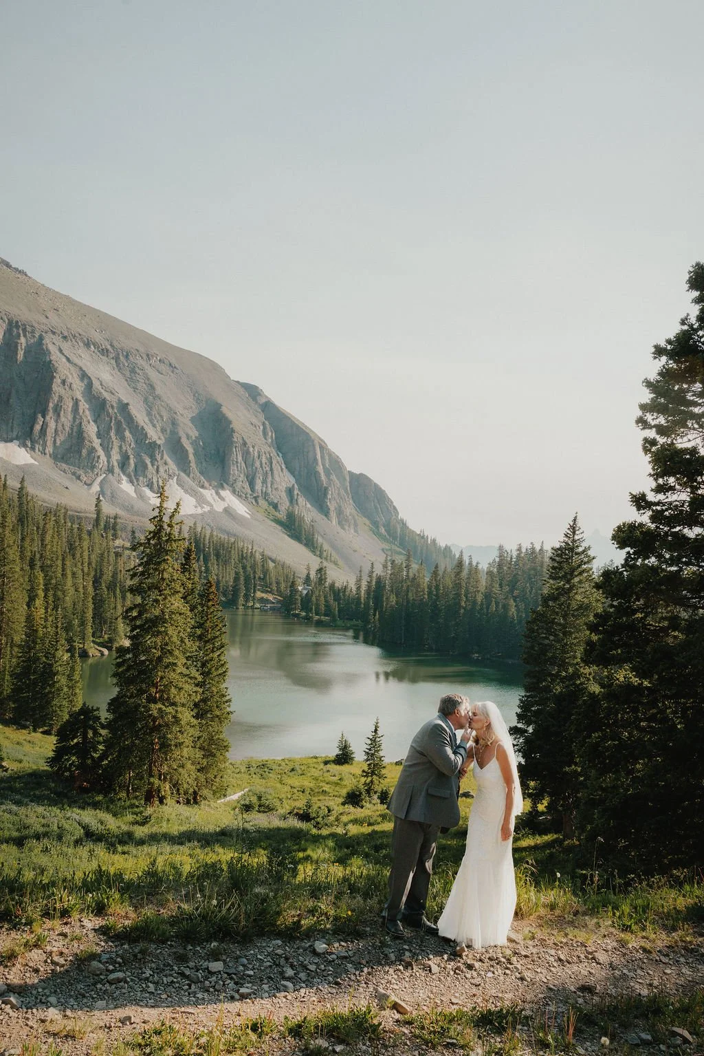 A wedding couple kissing outdoors in a mountainous forest landscape with a lake, trees, and mountains in the background.