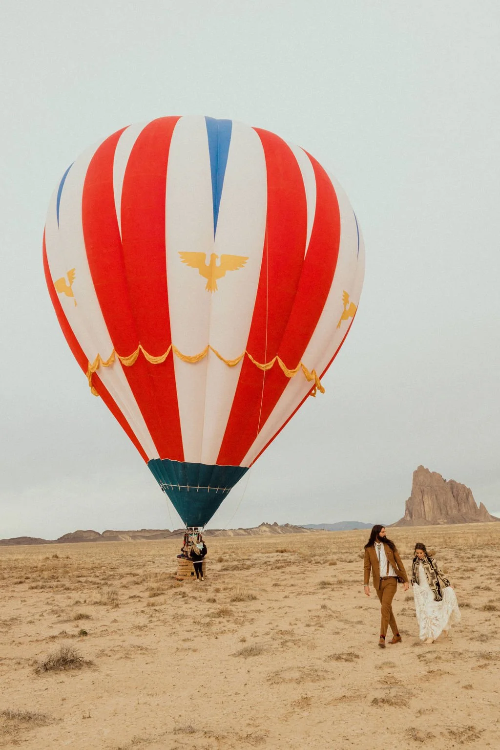 A hot air balloon with red, white, and blue vertical stripes and gold eagle symbols floats over a desert landscape with rocky formations in the background. Two women walk on the sandy ground below, wearing masks and casual clothing.