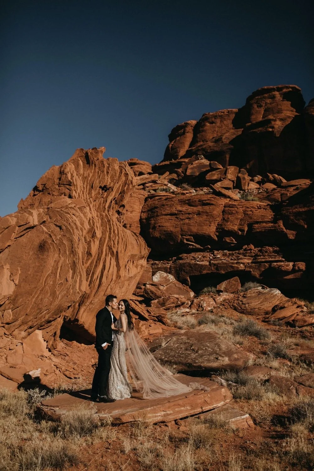 A bride and groom stand together in a desert landscape with red rocks and cliffs during twilight, the bride wearing a long veil and white gown, and the groom in a black suit.