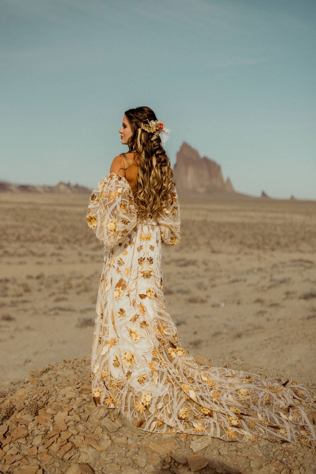 A woman in a long floral dress with puffy sleeves stands on rocky ground in a desert with a large rock formation in the background.