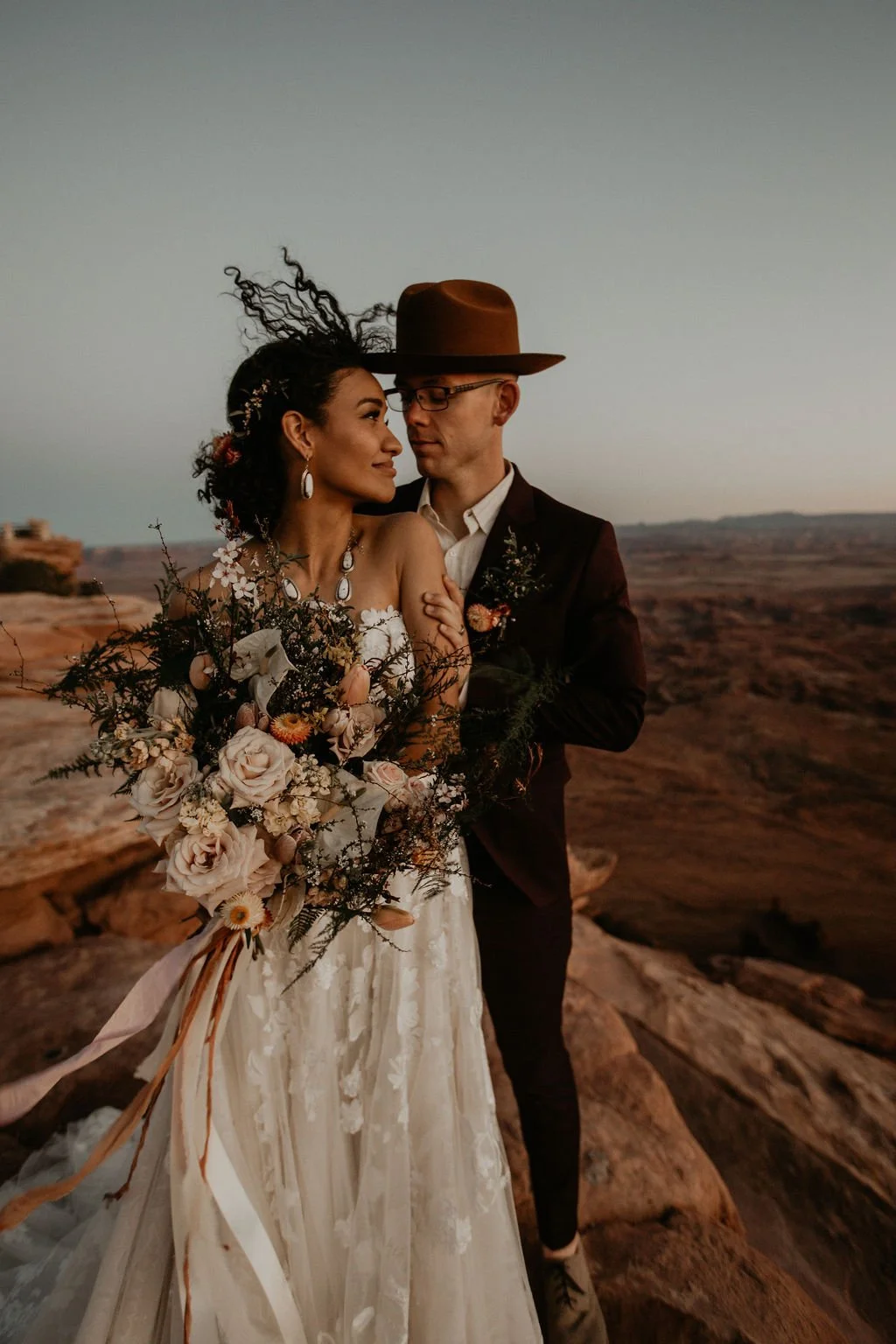 A couple dressed in wedding attire, embracing outdoors at sunset in a desert landscape, with the woman holding a large bouquet of flowers.