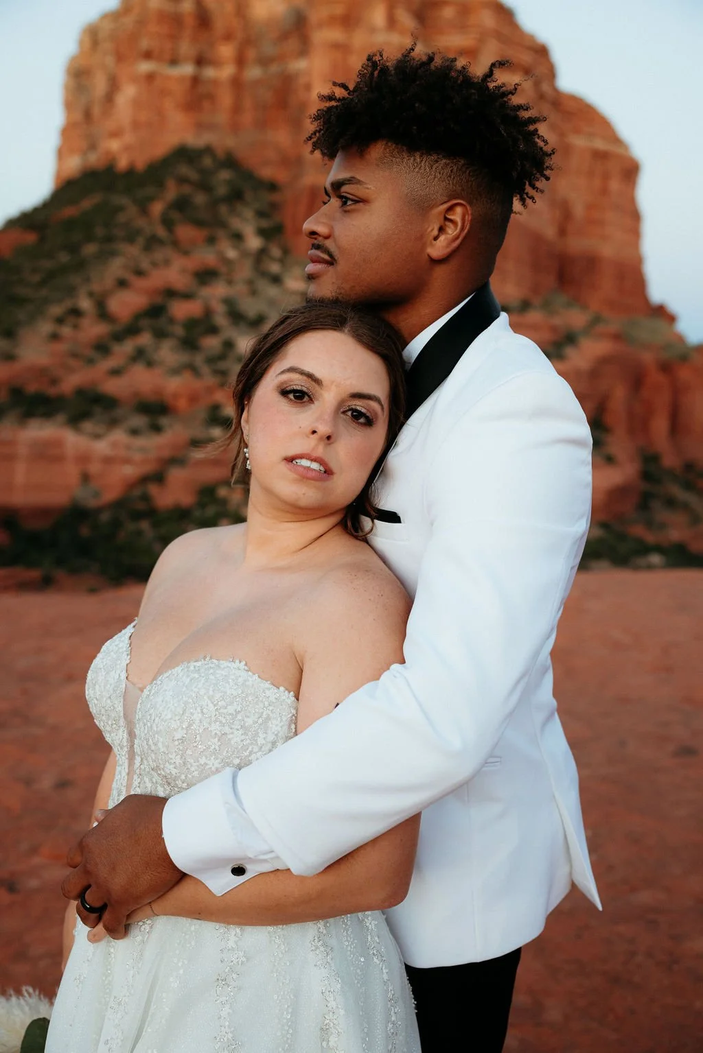 A couple dressed in wedding attire embracing outdoors with red rock formations in the background.