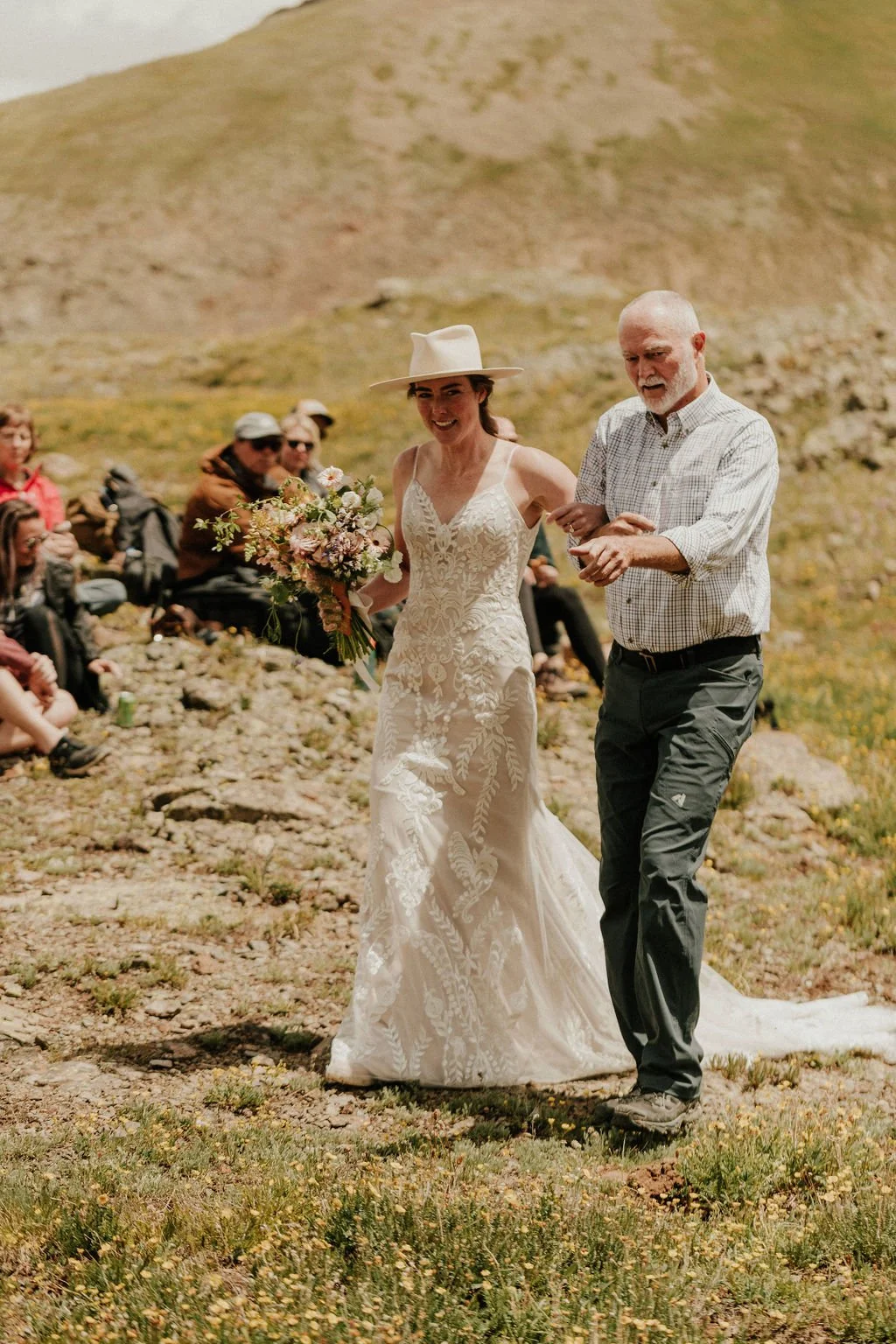 A bride in a white lace wedding dress holding a bouquet walks outdoors with an older man in a plaid shirt helping her.