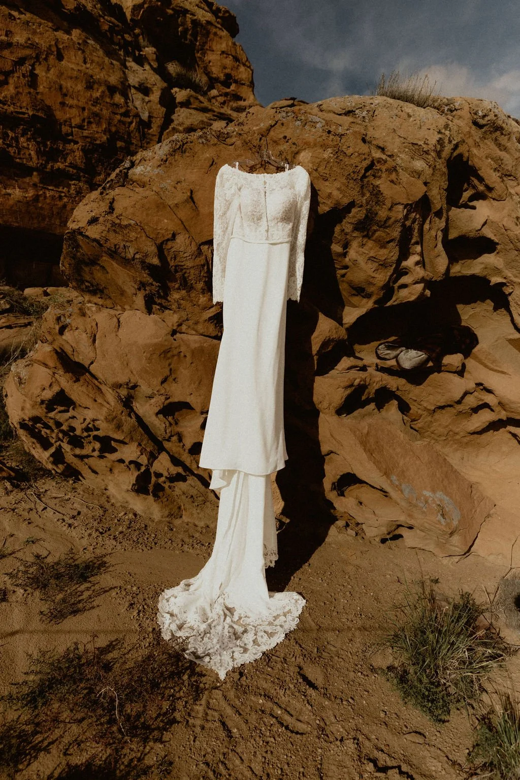 A white wedding dress hangs on a rock formation in a desert landscape with red rocks and sparse vegetation under a cloudy sky.