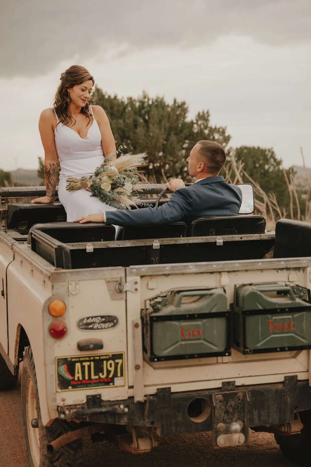 A couple in wedding attire sitting in the back of a vintage Land Rover vehicle outdoors under cloudy skies, with trees and rustic wooden structures in the background.